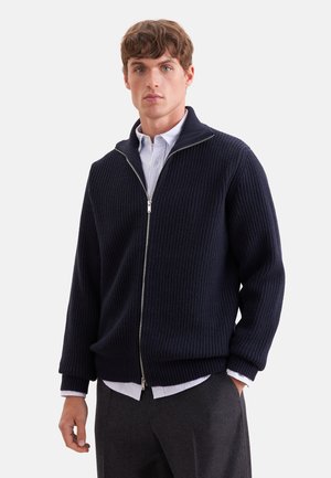 Ung mand iført navyblå ribbet sweat-shirt med lynlås over en hvid skjorte med krave og mørkegrå bukser stående mod en ensfarvet baggrund.