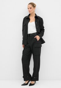 Zwarte op maat gemaakte blazer met twee voorzakken, gecombineerd met bijpassende cargobroek met zijzakken. Model draagt een witte top en zwarte hakken.