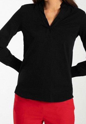Vrouw draagt een zwart shirt met lange mouwen en een V-hals en felrode broek, handen rustend op de heupen, torso zichtbaar.