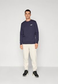 Felpa blu navy in misto cotone con polsini a costine, abbinata a pantaloni color crema. Il modello indossa scarpe da ginnastica nere.