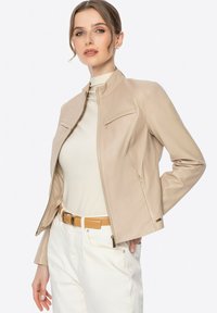Veste en cuir beige au design ajusté, avec un col montant et deux poches zippées sur la poitrine. Portée par-dessus un haut clair et un pantalon blanc.