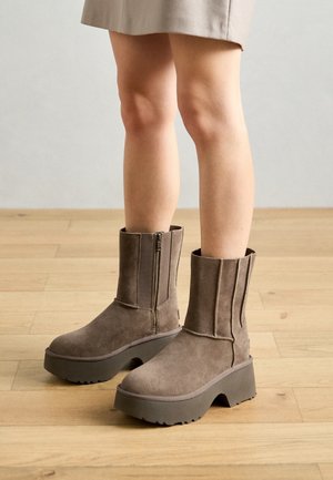 Jambes portant des bottines compensées en suède taupe avec fermetures éclair latérales, posées sur un sol en bois clair, jupe beige visible au-dessus des genoux.