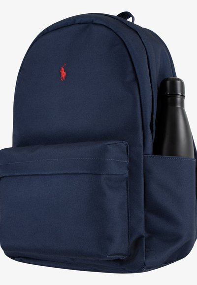 Ryggsäck i marinblått tyg med rundat lock, synligt framfack och en svart vattenflaska i sidofacket. Röd logotypdetalj.