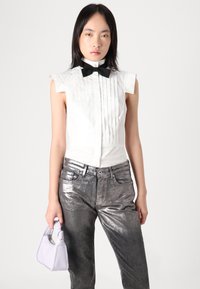 Blusa blanca plisada con un cuello alto y una pajarita negra, combinada con jeans plateados brillantes y un pequeño bolso de lavanda.