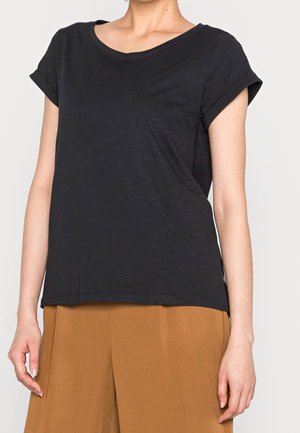 Kobieta ubrana w gładki czarny t-shirt z krótkim rękawem z podwiniętymi mankietami i brązowe spodnie z wysokim stanem, stojąca na jednolitym tle.