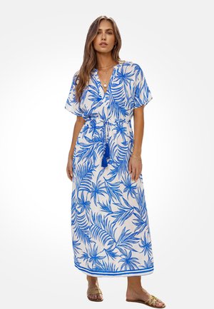 Vrouw draagt een witte maxi-jurk met blauwe tropische bladeren, korte mouwen, een strik in de taille, gelaagde kettingen en gouden sandalen.