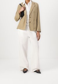 Blazer di lino beige con due tasche frontali, abbinato a pantaloni bianchi a gamba larga e scarpe nere con punta appuntita, mentre tiene una borsa nera.