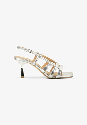Sandale argentée à brides ouvertes, talon kitten bas et bride de cheville ajustable sur fond blanc.