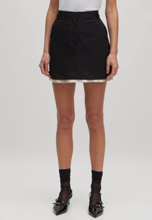 Zwarte rok met een gladde textuur, heeft een witte satijnen rand aan de zoom, een aansluitende taille en een recht model. De benen zijn blote en dragen donkere sokken.