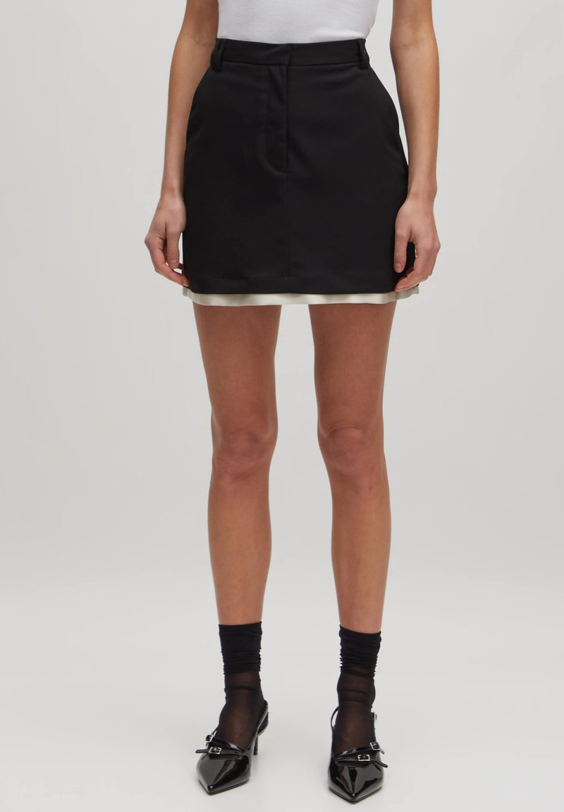 Zwarte rok met een gladde textuur, heeft een witte satijnen rand aan de zoom, een aansluitende taille en een recht model. De benen zijn blote en dragen donkere sokken.
