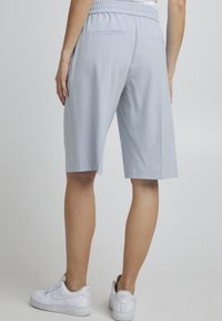 Osoba v světle modrých kraťasech do kolen s elastickým pasem a bílými teniskami, stojící na jednobarevném světle pozadí.