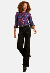 Krāsains rakstains blūze purpura un rozā toņos, kopā ar melnām augsto jostasvietu platām džinsām un melnām augstpapēžu kurpēm.