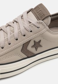 Beige leren sneaker met een gestructureerd bovenwerk, bijpassende veters en een bruin sterrenlogo. Platte zool met witte en zwarte streepaccenten.