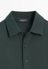 Dunkelgrünes Poloshirt aus strukturiertem Stoff, mit zwei schwarzen Knöpfen und einem klassischen Kragen. Das Etikett trägt den Aufdruck "Massimo Dutti."