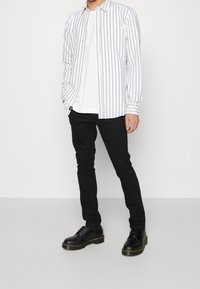 Gestreept overhemd met lange mouwen in wit en grijs, gelaagd over een wit T-shirt, gecombineerd met zwarte slim-fit broek en zwarte veterschoenen.