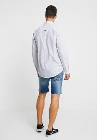 Muž oblečený v bílo-modrém proužkovaném triku s dlouhým rukávem, džínových šortkách a černých teniskách, stojící zády k fotoaparátu na jednoduchém pozadí.