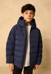 Garçon portant une doudoune bleu marine avec capuche sur une chemise blanche, se tenant devant un fond beige uni.
