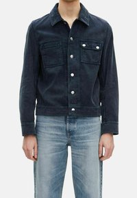 Man med en mörkblå kordjacka med silverknappar och två bröstfickor, ihop med ljusblå blekta jeans.