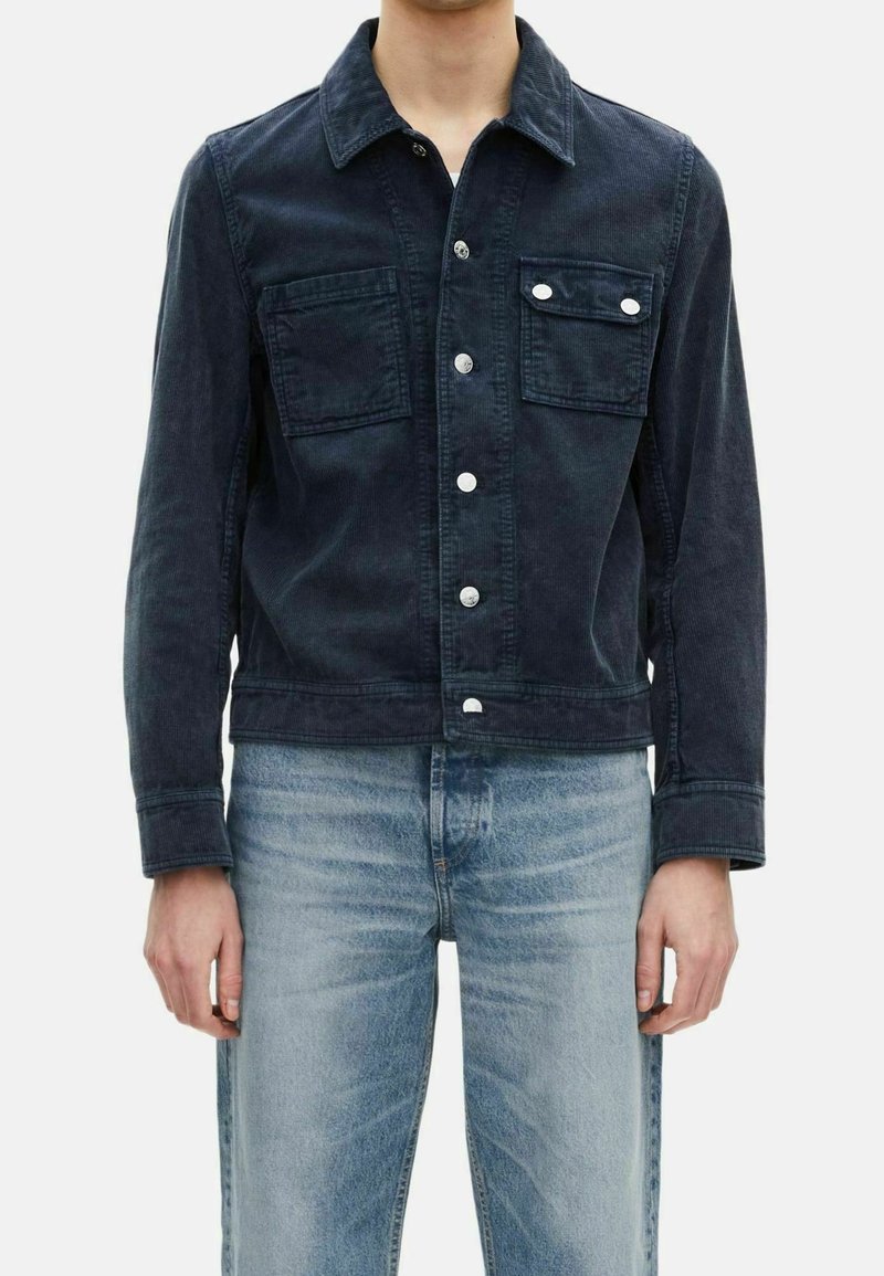 Man med en mörkblå kordjacka med silverknappar och två bröstfickor, ihop med ljusblå blekta jeans.