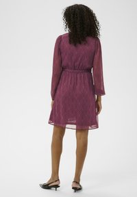 Lilla lühike kleit, millel on lehepilt, poolläbipaistev kangas, pikad varrukad ja hästi määratletud vöökoht, mille küljes on nöör. Kandmise all on mustade kontsakingadega.