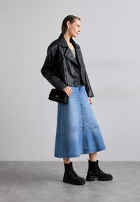 Veste en cuir synthétique noire sur un haut gris, associée à une jupe en denim bleu clair. Des bottines noires ornées et un petit sac à main noir complètent le look.