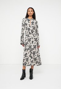 Robe longue beige avec un motif floral noir, manches longues et encolure en keyhole. Associée à des bottines noires.
