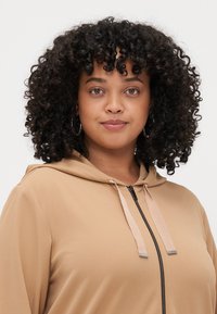 Μπεζ φούτερ με κουκούλα, με λεία υφή, μπροστινό φερμουάρ και αντίθετα κορδόνια. Διαθέτει χαλαρή εφαρμογή και απλό σχέδιο.