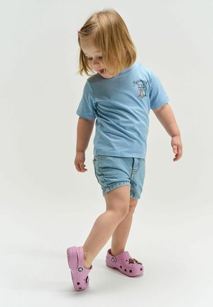 Fetiță mică cu tricou albastru deschis cu textul „Sirena”, pantaloni scurți din denim și Crocs roz, uitându-se în jos în timp ce face un pas înainte pe un fundal alb.