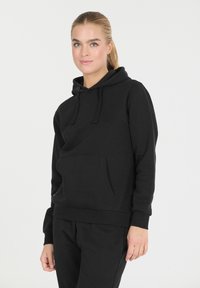 Nuori nainen, jolla on vaaleat hiukset, pukeutuneena mustaan huppariin ja mustiin housuihin, seisoo tasaisella valkoisella taustalla.