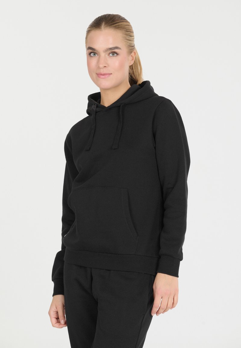 Nuori nainen, jolla on vaaleat hiukset, pukeutuneena mustaan huppariin ja mustiin housuihin, seisoo tasaisella valkoisella taustalla.