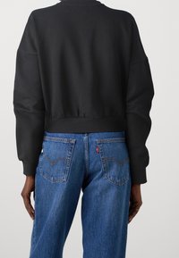 Personne portant un sweat-shirt court noir à manches longues et un jean en denim bleu vue de dos sur un fond uni.