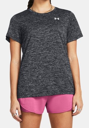 Frau trägt ein meliertes dunkelgraues sportliches Kurzarmshirt und leuchtend pinke Laufshorts, mit Schmuck an Händen und Hals.