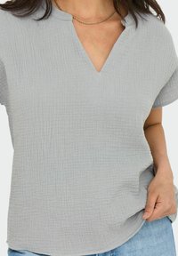 Top grigio chiaro, testurizzato, con scollatura a V, maniche corte e un sottile motivo a griglia. Tessuto morbido che si adagia delicatamente sul corpo.