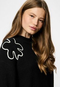 Pull en maille noire avec un motif floral blanc texturé sur l'épaule gauche ; matière douce avec une coupe décontractée et un col montant.