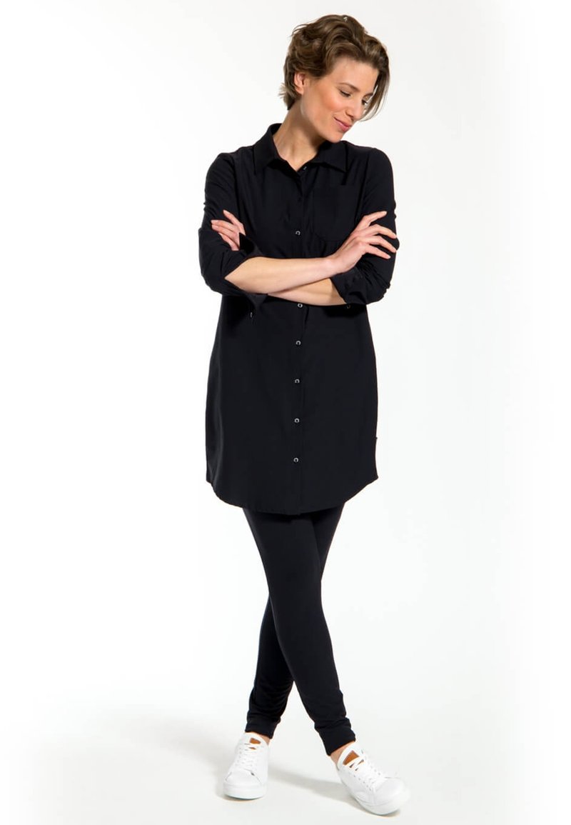 Zwarte lange mouwen knoopsluiting shirt met een gebogen onderkant, gecombineerd met zwarte leggings en witte sneakers. Gladde stof, klassieke kraag, enkele borstzak.