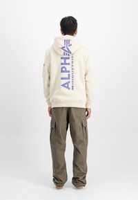 Krēmkrāsas kapuce ar purpursarkano "ALPHA" logotipu aizmugurē, savienota ar brīviem olīvu krāsas cargo biksēm ar sānu kabatām. Gluda tekstūra.