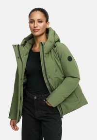 Groene pufferjack met een grote capuchon, ritssluiting, drukknopen en een ronde logo-patch op de linker mouw. Draagt over een zwarte top.