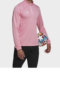 Rosa långärmad träningsoverdel med kvartdragkedja, med ribbade muddar, bärs med svarta byxor, och håller en flerfärgad fotboll.