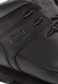 Bota de senderismo de cuero negro con tobillo acolchado, cordones negros y un acabado texturizado. El logo de Timberland se muestra de manera prominente.