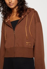 Hnědá krátká mikina na zip z měkké látky, s kapucí, zlatými detaily a logem Nike s nápisem "AIR" ve zlaté výšivce.