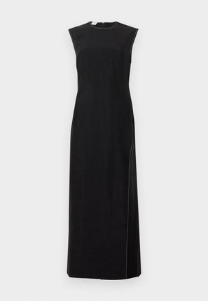 Ψηλή γυναίκα που φορά μακρύ μαύρο φόρεμα χωρίς μανίκια με πλαϊνή σκισμή και μαύρα μυτερά παπούτσια, στέκεται μπροστά σε απλό λευκό φόντο.