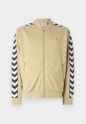 Veste bomber beige avec un motif subtil, rayures chevron navy sur les manches, fermeture éclair et poches latérales. Présente un logo sur la poitrine.