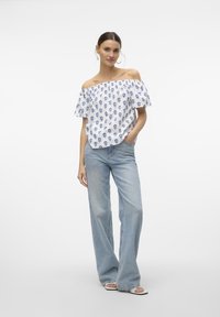 Hvid off-the-shoulder top med blåt kraniummønster, bløde ærmer, sat sammen med lyseblå højtaljede jeans og hvide sandaler.