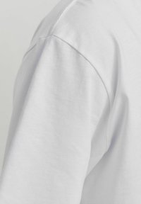 Detalle de una camisa blanca de manga corta que muestra una textura suave y lisa, con una costura de hombro redondeada y un sutil bordado a lo largo del escote.