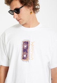 Hvid bomulds t-shirt med et farverigt grafisk design med øjne indeholdt i et lilla rektangel og orange detaljer. Korte ærmer, rund halsudskæring.