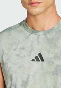 Jasnozielona, bezrękawnikowa koszulka sportowa z teksturowanym wzorem i czarnym logo Adidas na piersi. Okrągły dekolt, oddychający materiał.