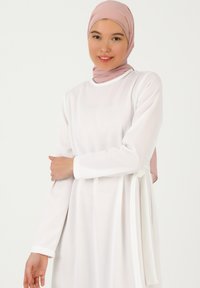 Long-sleeved white dress with a relaxed fit, featuring a tie at the waist. Paired with a light pink scarf, accentuating the overall look.