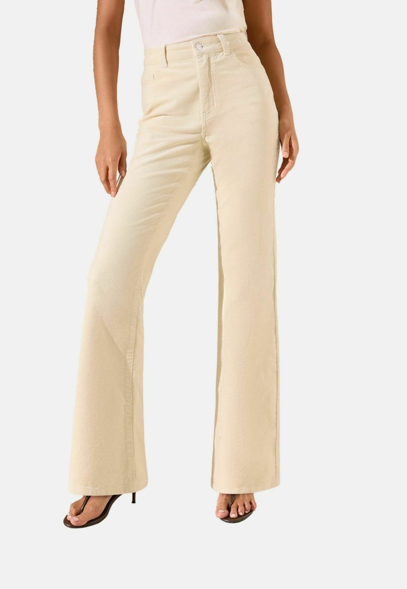 Pantalon évasé beige en tissu texturé, avec une taille haute, une poche avant et des détails de couture subtils. Associé à des sandales à talons noires.