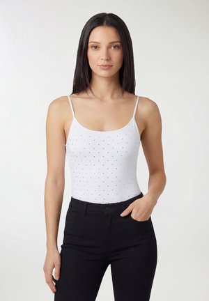 Kobieta o długich ciemnych włosach, ubrana w biały bezrękawnik w małe kropki i czarne spodnie, stojąca z jedną ręką w kieszeni.