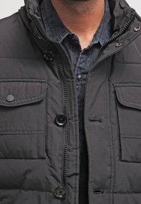 Czarna pikowana kurtka z suwakiem z przodu, dwoma kieszeniami na piersi z klapkami na guziki oraz kapturem. Gładka faktura i casualowy design.